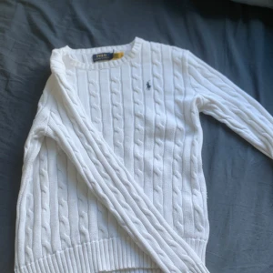 Vit kabelstickad tröja från Polo Ralph Lauren - Säljer en klassisk vit kabelstickad tröja från Polo Ralph Lauren med rund halsringning och broderad logga på bröstet. Tröjan har lång ärm och ribbade muddar vid ärmslut och nederkant. Perfekt till många olika outfits.