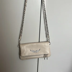 Beige axelväska med kedjerem och silverdetaljer - Säljer en beige axelväska i skinn med silverfärgad kedjerem och dekorativt emblem framtill. Väskan har dubbla dragkedjor på framsidan och ett extra fack med dragkedja på baksidan. Perfekt storlek för det viktigaste.EJ EKTA