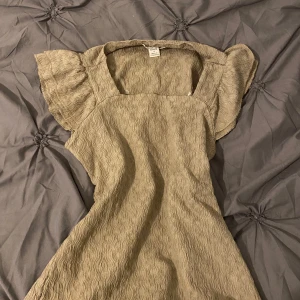 Beige topp med struktur och volangärmar - Säljer en beige topp med fyrkantig ringning och korta volangärmar. Toppen har en strukturerad yta och en feminin siluett som passar perfekt till vår och sommar. Superfin att styla med sneakers eller sandaler!