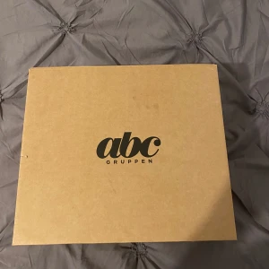 Studentmössa från abc Gruppen - Klassisk studentmössa med vit ovandel, svart band och blank skärm. Insidan är fodrad med gul satin och har märkning från abc Gruppen. Framtill sitter en gul dekoration med blå detalj.