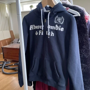 Mörkblå hoodie från Abercrombie & Fitch - Mörkblå hoodie från Abercrombie & Fitch med vit broderad logga och text framtill. Tröjan har dragkedja, huva med vita snören och två fickor fram. Perfekt för en avslappnad stil. Stl M