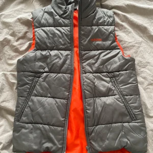 Grå väst från Adidas - Snygg grå väst från Adidas med orange insida och dragkedja framtill. Västen har två fickor och en hög krage. Osäker på storlek men passar XS/S