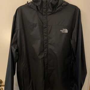 Svart vindjacka från The North Face - Säljer en svart vindjacka från The North Face med huva och dragkedja. Jackan har en stilren design med logga på bröstet och är perfekt för blåsiga dagar. Passar dig som vill ha en lätt och funktionell jacka.