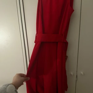 Röd ärmlös jumpsuit med bälte - Säljer en snygg röd jumpsuit med bälte i midjan. den  är ärmlös och har en elegant, rak passform som ger en stilren look. Perfekt för dig som vill sticka ut med en färgstark outfit.