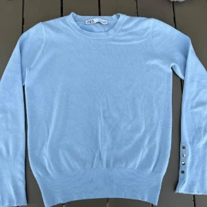 Ljusblå tröja från Zara - Säljer en ljusblå långärmad tröja från Zara med rund halsringning. Tröjan har ribbade muddar och dekorativa knappar vid ärmslutet på ena ärmen. Perfekt för en stilren look.