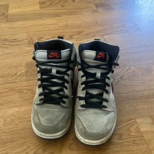 Nike Dunk High grå med svart och röd detalj - Säljer ett par Nike Dunk High sneakers i grått med svart swoosh och röda detaljer. Skorna har svart snörning och hög modell som ger en klassisk streetwear-look. Perfekta för dig som gillar stilrena och sportiga sneakers.