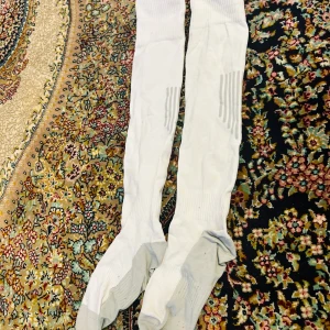 Vita knälånga sportstrumpor - Säljer ett par vita knälånga sportstrumpor med grå förstärkning vid tår och häl. Den har ribbade partier för extra stöd och sitter uppe bra vid aktivitet. Perfekta för träning eller fotboll.