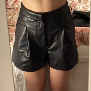 Svarta shorts i skinnimitation - Snygga svarta shorts i skinnimitation med hög midja och veck framtill. Perfekta för att lyfta vilken outfit som helst. Stängs med dragkedja i sidan och har en stilren look.