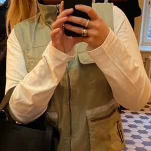 Beige väst med reflexdetaljer och fickor - Beige väst med flera praktiska fickor och reflexdetaljer framtill. Västen har dragkedja och hög krage, perfekt för utomhusaktiviteter. Tillverkad i ett lätt syntetmaterial.