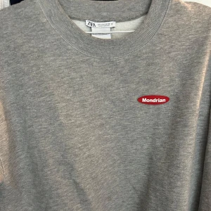 Grå sweatshirt med Mondrian-tryck från Zara - Grå sweatshirt från Zara med Mondrian-tryck på ryggen och liten röd Mondrian-logga framtill. Klassisk rund halsringning och mjukt material.