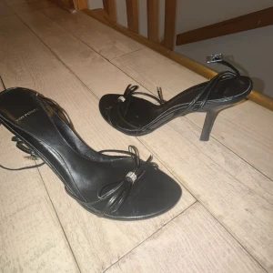 Svarta högklackade sandaler med smala remmar - Säljer ett par svarta sandaler med hög klack och smala remmar. Skorna har öppen tå, dekorativ knut framtill och ett litet spänne runt vristen. Perfekta för dig som gillar stilrena och eleganta skor.❤️