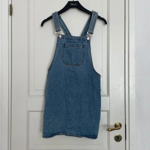 Jeansklänning med hängslen - Säljer en klassisk jeansklänning med hängslen och ficka framtill. Perfekt att styla med en t-shirt eller tröja under för en avslappnad look. Klänningen har justerbara axelband och är i en tidlös blå denim.