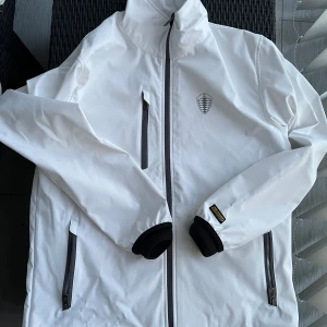 Vit vindjacka från Blåkläder - Säljer en vit vindjacka från Blåkläder med svarta detaljer och dragkedjor. Jackan har hög krage, långa ärmar och TEAM Koenigsegg-tryck på ryggen. Perfekt för dig som gillar stilren och sportig design.