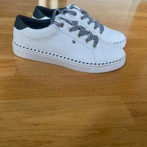 Vita sneakers från Tommy Hilfiger - Snygga vita sneakers från Tommy Hilfiger med svart detalj vid hälen och randiga svarta/vita skosnören. Skorna har diskret logga på sidan och dekorativ rand längs sulan och även loggan på flärpen. Perfekta för en stilren och sportig look. Storlek 39. Endast använda en gång