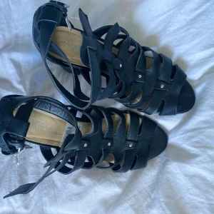 Svarta sandaler med klack från Vox Shoes - Säljer ett par svarta sandaler från Vox Shoes med bred klack och öppen tå. Skorna har flera remmar med små silvriga nitar och spänne runt vristen. Perfekta för dig som vill ha en stilren och trendig look.