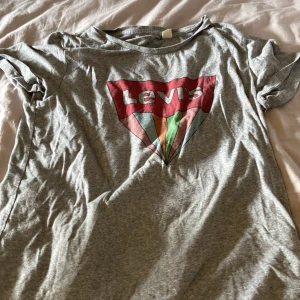 Grå t-shirt från Levi's med färgglad logga - Säljer en grå t-shirt från Levi's med en stor, färgglad logga på bröstet. T-shirten har rund halsringning och korta ärmar. Perfekt för en avslappnad stil. ca 80 cm i midjan
