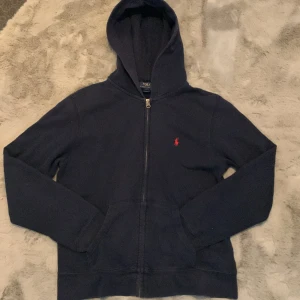 Mörkblå hoodie från Polo Ralph Lauren - Mörkblå hoodie med dragkedja från Polo Ralph Lauren. Klassisk design med huva, fickor framtill och röd broderad logga på bröstet. Perfekt för en avslappnad stil. Inga skadade delar och fräsch. Nytt pris 1100 kr mitt pris ändast 400 kr går att pruta 