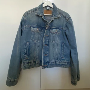 Blå jeansjacka  - Säljer en klassisk blå jeansjacka med knappar framtill och två bröstfickor med lock. Jackan har en rak, normal passform. 