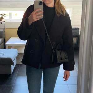 Svart kappa med dubbelknäppning - Säljer en svart kappa med dubbelknäppning och klassisk krage. Jackan har lång ärm och en stilren, tidlös design som passar perfekt till jeans eller kostymbyxor. Enkel att matcha och snygg till vardagslooken.