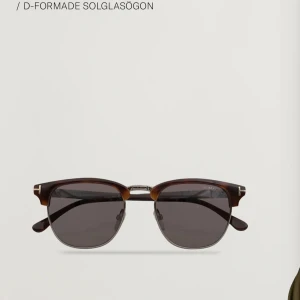 Bruna D-formade solglasögon från Tom Ford - Snygga solglasögon från Tom Ford med D-formade bågar i brun och svart. Glasen är mörka och bågarna har en klassisk clubmaster-stil med metallbrygga. Perfekt accessoar för en stilren look.