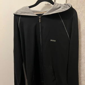  hoodie  BOSS - Svart hoodiejacka från BOSS med grå huva och dragkedja framtill. Jackan har fickor på magen och diskret BOSS-logga på bröstet. Perfekt för en avslappnad stil.