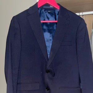 Mörkblå kavaj från Riley - Stilren mörkblå kavaj från Riley med två knappar framtill och klassiska fickor. Perfekt för bal, student eller fest. Tunnare foder och enkel design som passar till många tillfällen. Den är köpt på brothers, från märket Riley. Den är som ny, och är perfekt för dig med storlek 46. Är generös med priset då den blivit för liten för mig. Den är köpt tillsammans med en kavaj, för 2000kr.