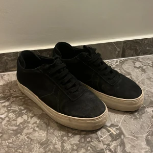 Svarta sneakers från Axel Arigato - Säljer ett par svarta sneakers från Axel Arigato med tjock vit sula och klassisk snörning. Skorna har en rund tå och är tillverkade i mocka, vilket ger en stilren och modern look. Perfekta för dig som gillar minimalistisk design.