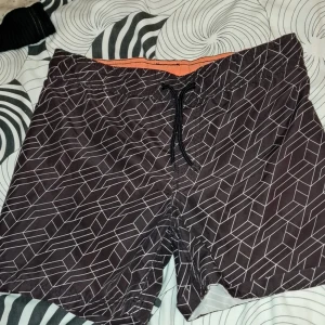 Svarta mönstrade badshorts från Lindex - Svarta badshorts med vitt geometriskt mönster och orange insida från Lindex. Shortsen har snörning i midjan och är perfekta för sommardagar vid vattnet.