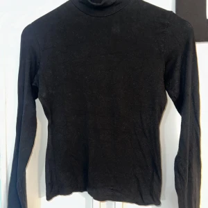 Svart polotröja i bomull - Säljer en klassisk svart polotröja med lång ärm. Tröjan är i mjukt bomullsmaterial och har en tight passform som sitter snyggt mot kroppen. Perfekt att styla med jeans eller kjol för en enkel och stilren look. Finns ett litet litet hål på underarmen på vänster arm. Syns endast om man sträcker upp armen och den sitter åt tajt just där. Annars syns den inte 