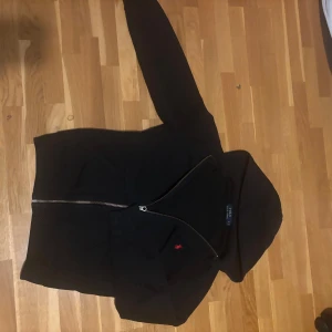 Svart hoodie från Polo Ralph Lauren - Säljer en svart hoodie från Polo Ralph Lauren med dragkedja och huva. Tröjan har en liten röd logga broderad på bröstet och två fickor framtill. Perfekt för en avslappnad stil.