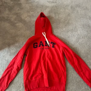 Röd hoodie från GANT - Säljer en röd hoodie från GANT i storlek M. Tröjan har huva med vita snören och tryckt GANT-logga samt texten 'Est. 1949' på bröstet. 
