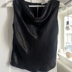 Svart satinlinne med draperad hals - Säljer ett svart linne i glansigt satinmaterial med snygg draperad halsringning. Toppen är ärmlös och har en enkel knäppning bak i nacken. Perfekt för att lyfta vilken outfit som helst. Var för liten för mig så är aldrig använd. Skriv för fler bilder💞