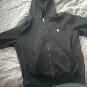 Svart hoodie med dragkedja från Ralph Lauren - Svart hoodie med dragkedja från Ralph Lauren. Klassisk design med huva, fickor framtill och den ikoniska lilla loggan broderad på bröstet. Perfekt för en avslappnad stil.