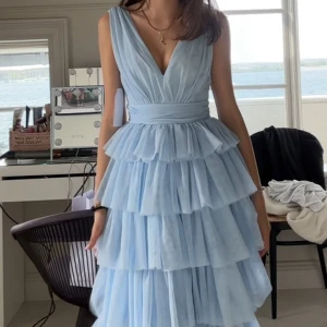 Ljusblå balklänning med volanger - Säljer en drömmig ljusblå långklänning med flera lager volanger och djup v-ringning. Klänningen har bred midja och är ärmlös, perfekt för bal eller fest. Materialet är lätt och ger ett svävande intryck. Som aldrig har använts 