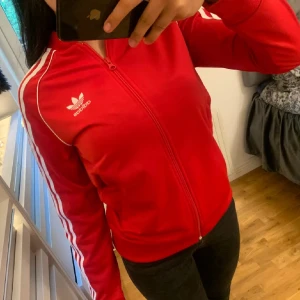 Röd Adidas kofta med vita ränder - En klassisk röd Adidas kota med vita ränder längs ärmarna och vit logga på bröstet. Jackan har dragkedja framtill, ståkrage och ribbade muddar. Storlek 158 men passar mig som bär Xs.