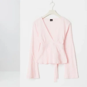 Ljusrosa omlottopp med knytband, ny med prislapp  - Supersöt ljusrosa omlottopp med v-ringning och långa ärmar. Toppen har knytband i midjan som knyts baktill för en snygg passform. Perfekt att styla med jeans eller kjol för en trendig look.