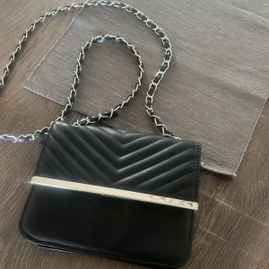 Svart axelväska med kedjerem - Snygg svart axelväska med quiltad ovandel och silverfärgad metallist framtill. Kedjerem med inslag av svart material ger en cool touch. Perfekt för att lyfta din outfit.