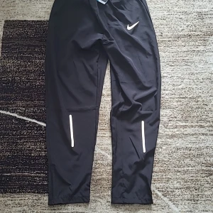 Svarta träningsbyxor från Nike Dri-Fit - Svarta träningsbyxor från Nike Dri-Fit med vit logga på ena benet och reflexdetaljer framtill. Byxorna har elastisk midja med dragsko och är tillverkade i ett lätt syntetmaterial. Perfekta för löpning eller gym.