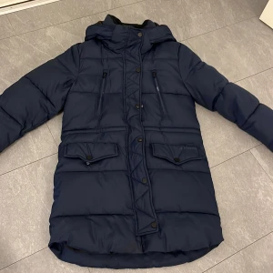 Mörkblå lång dunjacka med huva - Säljer en mörkblå lång dunjacka med huva och flera fickor med både dragkedja och knappar. Jackan har en pufferstil och är perfekt för kalla dagar. Reflextape baktill för extra synlighet. Stängs med dragkedja och knappar framtill.