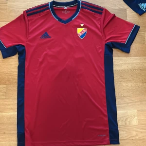 Röd Djurgården fotbollströja från Adidas - Säljer en röd Djurgården fotbollströja från Adidas med blå detaljer och klubbmärke på bröstet. Tröjan har korta ärmar, blå ränder på axlarna och nummer 19 på ryggen. Perfekt för match eller träning!