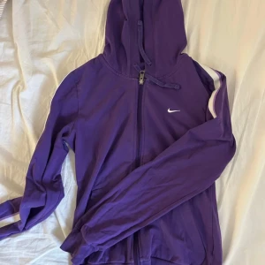 Lila hoodiejacka från Nike - Säljer en lila hoodiejacka från Nike med dragkedja och huva. Jackan har vita ränder längs ärmarna och en liten vit Nike-logga på bröstet. 