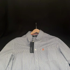 Rutig skjorta från Sail Racing - Snygg blå- och vitrutig skjorta från Sail Racing med klassisk krage och knappar framtill. Skjortan har en liten orange logga broderad på bröstet och detaljer i orange på ryggen. Perfekt för dig som gillar stilrena och sportiga plagg. Ny pris 1200kr