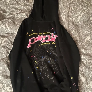 Svart hoodie från SP5DER med stjärnor och spindelnätsmönster - Cool svart hoodie från SP5DER med stort tryck framtill i rosa och vitt, text och spindelnätsmönster samt färgglada stjärnor. Baksidan har en gul stjärna och ärmarna är dekorerade med små stjärnor. Perfekt statement-plagg!