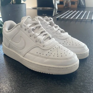 Vita Nike Court Vision Low  - Klassiska helvita Nike Court Vision Low sneakers med lågt skaft och perforerade detaljer på tån. Skorna har snörning och är tillverkade i riktigt skinn och syntetskinn med en platt sula. Perfekta för en clean och stilren look.