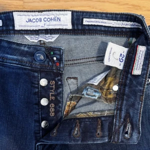 Jacob Cohën mörkblå jeans Style 688 - Säljer ett par mörkblå jeans från Jacob Cohën, modell Style 688. Klassisk femficksdesign med raka ben och snygga detaljer vid knapparna. Jeansen har en stilren look och är tillverkade i ett mjukt denimtyg.