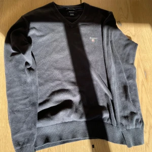 Mörkgrå v-ringad tröja från GANT - Säljer en mörkblå v-ringad tröja från GANT i mjuk bomull. Tröjan har långa ärmar, ribbade muddar och en liten broderad logga på bröstet. Perfekt för en stilren och klassisk look.