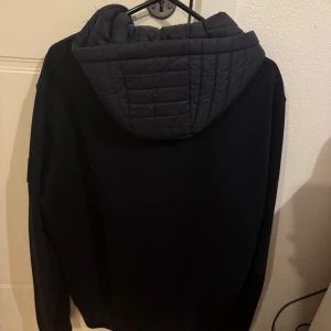 Svart hybridjacka med huva från Tommy Hilfiger - Snygg svart jacka från Tommy Hilfiger med quiltad framsida och huva. Jackan har dragkedja framtill och en patch med logga på ärmen. Perfekt för dig som vill ha en stilren och sportig look.