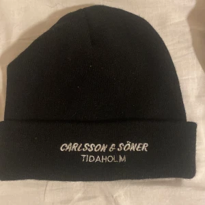 Svart stickad mössa med broderad text - Svart, klassisk stickad mössa från Carlsson & Söner med broderad vit text 'TIDAHOLM' framtill. Perfekt för kalla dagar och enkel att matcha med olika outfits.