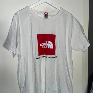 Vit t-shirt från The North Face - Vit t-shirt från The North Face med rött logotryck på bröstet och liten svart logga på ryggen. Klassisk passform och rund halsringning. Perfekt för en avslappnad stil.