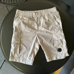 Silver shorts från C.P. Company - Säljer ett par silver cargoshorts från C.P. Company med elastisk midja och dragsko. Shortsen har flera fickor, inklusive två stora cargofickor på sidorna och en bakficka. Klassisk C.P. Company-linsdetalj på ena benet. Skick som ny, använd bara en gång. Ingå hål eller fläckar. Färg är silver, reflekterar mot ljus och blixter lite i mörket. Väldigt snygga och fräscha shorts som är givetvis äkta. Vid frågor och funderingar skriv pm. OBS Skickas ej! Bara avhämtning i Göteborgsområdet/ Mölndal